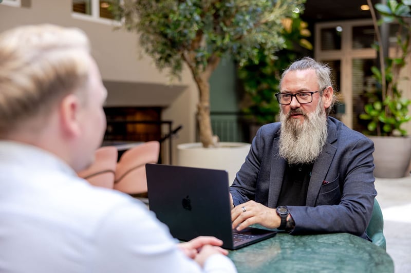 A man with glasses and a long beard working on a laptop across the table from a colleague