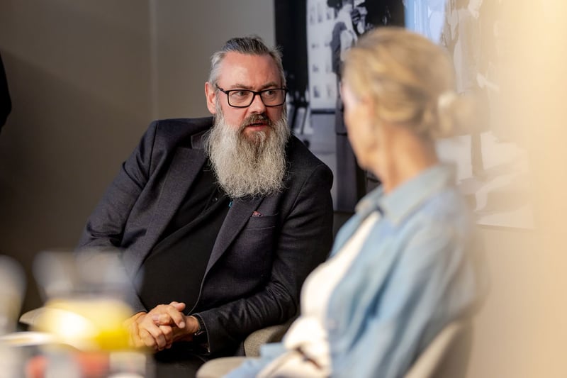 A man with glasses and a long beard listening in a conversation with a colleague